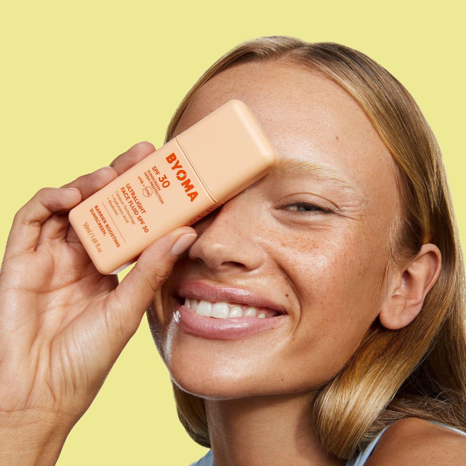 Smiling woman holding a bottle of BYOMA SPF 30 sunscreen to her face.