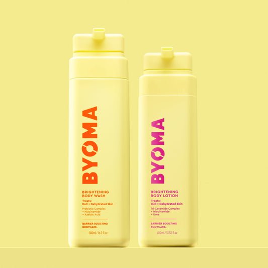 Two yellow BYOMA bottles on a pastel yellow background. Left: Brightening Body Wash with orange-red text; right: Brightening Body Lotion with pink text. Both are upright.