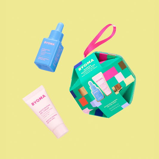 A green, multi-colored ornament-shaped gift box labeled “BYOMA Barrier Boosting Duo,” featuring a pink tube of moisturizing gel-cream and a blue bottle of hydrating serum. The items are shown floating against a yellow background.
